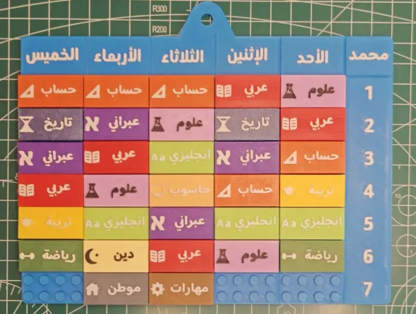 لوح جدول حصص أزرق مطبوع ثلاثي الأبعاد، عليه قطع ملوّنة للمواد واسم الطفل فوق عاليمين